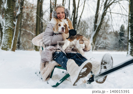 Young girl with blonde hair wearing winter clothing is sitting on a sled in the snow, holding a beagle dog while surrounded by trees in a winter landscape Young girl with blonde hair wearing winter clothing is sitting on a sled in the snow, holding a beagle dog while surrounded by trees in a winter landscape 140015391