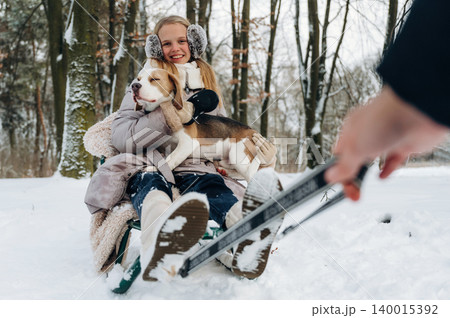 Young girl with long blonde hair wearing earmuffs is sitting on a sled in the snow, holding a beagle dog while a hand is pulling the sled in a winter forest setting Young girl with long blonde hair wearing earmuffs is sitting on a sled in the snow, holding a beagle dog while a hand is pulling the sled in a winter forest setting 140015392