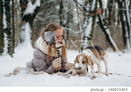 Young woman with blonde hair wearing earmuffs is playing with a beagle dog on a blanket in a snowy forest during winter season with trees in the background Young woman with blonde hair wearing earmuffs is playing with a beagle dog on a blanket in a snowy forest during winter season with trees in the background 140015403