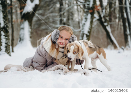 Young girl with blonde hair wearing earmuffs is lying in the snow beside a beagle dog, surrounded by snow-covered trees in a winter forest setting with a cozy blanket 140015408