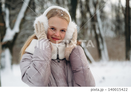 Young girl with long blonde hair wearing earmuffs and gloves smiles in a snowy forest, dressed in a warm coat during winter season with trees in the background 140015422