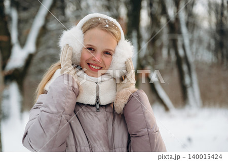 Young girl with long blonde hair wearing winter clothing and earmuffs smiles while standing in a snowy forest, surrounded by trees and a winter landscape with soft snowfall Young girl with long blonde hair wearing winter clothing and earmuffs smiles while standing in a snowy forest, surrounded by trees and a winter landscape with soft snowfall 140015424