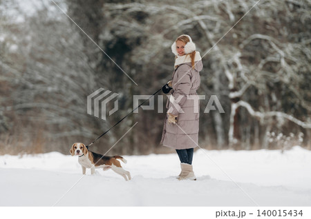 Woman in a long winter coat walking a beagle dog on a snowy landscape with trees in the background during a cold day in a serene outdoor setting Woman in a long winter coat walking a beagle dog on a snowy landscape with trees in the background during a cold day in a serene outdoor setting 140015434