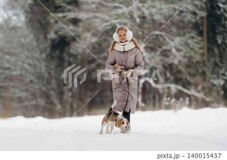 Young woman with long blonde hair wearing a warm winter coat walks a beagle dog on a snowy path surrounded by trees in a winter landscape with copy space 140015437
