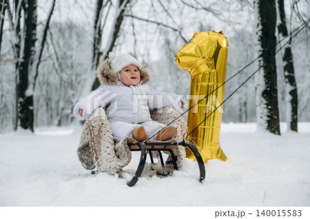 Baby in white winter outfit sitting on sled in snowy forest, holding golden number one balloon, celebrating first birthday with joyful atmosphere and winter wonderland backdrop 140015583