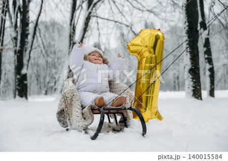 Baby in white snowsuit is joyfully seated on a sled in a snowy landscape, surrounded by trees, celebrating first birthday with a golden balloon number one 140015584