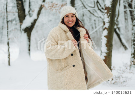 Smiling woman in cozy cream coat and knitted hat stands in snowy forest, surrounded by trees, showcasing winter fashion and joyful atmosphere in nature Smiling woman in cozy cream coat and knitted hat stands in snowy forest, surrounded by trees, showcasing winter fashion and joyful atmosphere in nature 140015621
