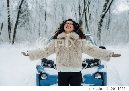 Young woman with curly hair wearing a fur coat stands joyfully in a snowy landscape with a blue car in the background surrounded by frosty trees and winter scenery Young woman with curly hair wearing a fur coat stands joyfully in a snowy landscape with a blue car in the background surrounded by frosty trees and winter scenery 140015631