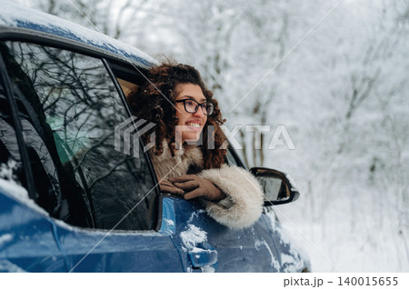 Woman with curly hair wearing a fur coat leans out of a blue car window, smiling in a snowy landscape with trees covered in white during winter season Woman with curly hair wearing a fur coat leans out of a blue car window, smiling in a snowy landscape with trees covered in white during winter season 140015655