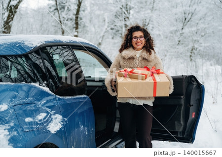 Woman with curly hair wearing a fur coat is holding a gift box with a red ribbon while standing next to a blue car in a snowy landscape with trees in the background Woman with curly hair wearing a fur coat is holding a gift box with a red ribbon while standing next to a blue car in a snowy landscape with trees in the background 140015667