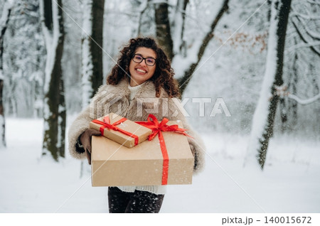 Young woman with curly hair wearing a fur coat is smiling while carrying wrapped gifts in a snowy forest during winter, surrounded by trees and falling snowflakes Young woman with curly hair wearing a fur coat is smiling while carrying wrapped gifts in a snowy forest during winter, surrounded by trees and falling snowflakes 140015672