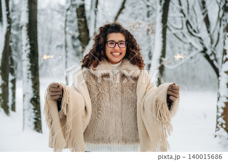 Smiling woman with curly hair, wearing a cozy beige shawl, holding sparklers in a snowy forest, celebrating winter festivities with joy and warmth 140015686
