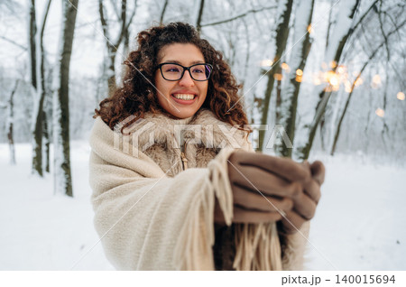 Smiling woman with curly hair wrapped in a cozy blanket stands in snowy forest, holding warm drink, surrounded by winter trees, creating a joyful holiday atmosphere 140015694