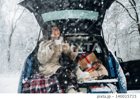 Woman enjoying a warm beverage while sitting in the back of a car, surrounded by snow-covered trees and holiday gifts, creating a cozy winter atmosphere with joy Woman enjoying a warm beverage while sitting in the back of a car, surrounded by snow-covered trees and holiday gifts, creating a cozy winter atmosphere with joy 140015715