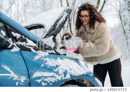 Woman with curly hair in a cozy fur jacket is pouring fluid into the engine of a blue car covered in snow, showcasing winter car maintenance and care 140015723