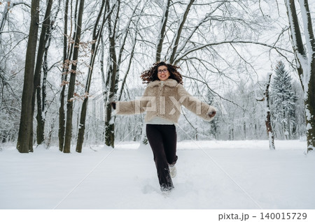 Young woman in a cozy fur jacket joyfully running through a snowy forest, surrounded by tall trees and a serene winter landscape, capturing the essence of winter fun and freedom 140015729