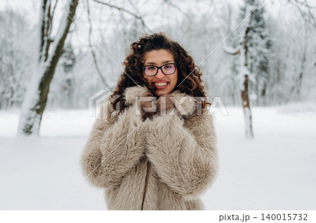 Young woman with curly hair, wearing a cozy fur coat, smiles warmly in a snowy forest, surrounded by trees, capturing the essence of winter joy and comfort Young woman with curly hair, wearing a cozy fur coat, smiles warmly in a snowy forest, surrounded by trees, capturing the essence of winter joy and comfort 140015732