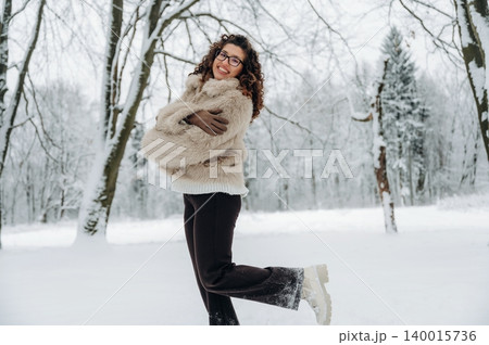 Smiling woman with curly hair wearing a cozy fur coat stands joyfully in a snowy landscape, surrounded by trees, capturing the essence of winter happiness and warmth 140015736