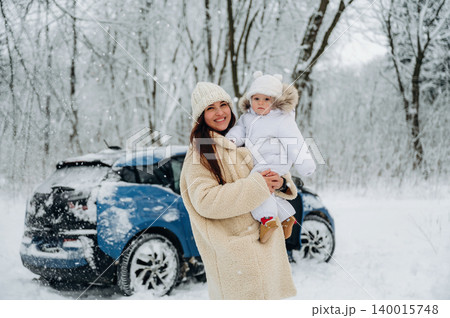 Woman holding child in winter attire, standing in snowy landscape with blue car in background, showcasing family bonding and winter adventure in a serene environment 140015748