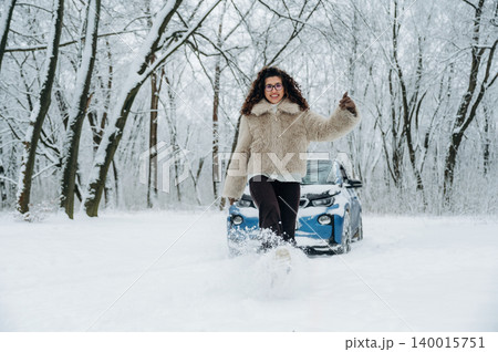 Young woman with curly hair wearing a fur coat is walking through a snowy forest with a blue car parked nearby, surrounded by white trees and winter scenery Young woman with curly hair wearing a fur coat is walking through a snowy forest with a blue car parked nearby, surrounded by white trees and winter scenery 140015751