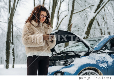 Young woman with curly hair wearing a fluffy coat is standing beside a blue car with an open hood in a snowy forest while checking her phone for assistance 140015764