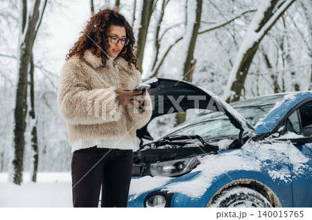 Young woman with curly hair wearing a fur coat is standing beside a blue car with an open hood in a snowy forest, checking her phone for assistance during a winter breakdown Young woman with curly hair wearing a fur coat is standing beside a blue car with an open hood in a snowy forest, checking her phone for assistance during a winter breakdown 140015765