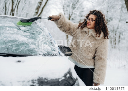 Woman with curly hair in a fluffy coat is clearing snow from a car windshield using a brush in a snowy outdoor setting surrounded by trees 140015775
