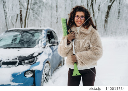 Young woman with curly hair wearing a fluffy coat is holding a snow shovel beside a car covered in snow in a winter forest setting with trees in the background Young woman with curly hair wearing a fluffy coat is holding a snow shovel beside a car covered in snow in a winter forest setting with trees in the background 140015784