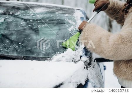 Woman using a green ice scraper to remove snow from the windshield of a car parked in a snowy environment during winter weather conditions 140015788