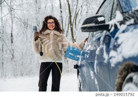 Woman with curly hair wearing a fur coat is charging an electric car in a snowy landscape, holding a charging cable while standing beside the vehicle in winter conditions 140015792