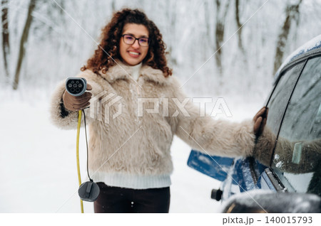 Woman with curly hair wearing a fluffy coat is holding an electric car charging cable near a vehicle in a snowy outdoor setting with trees in the background 140015793