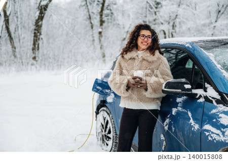 Young woman with curly hair wearing a fur coat is standing beside an electric car charging in a snowy landscape, holding a warm drink during winter 140015808