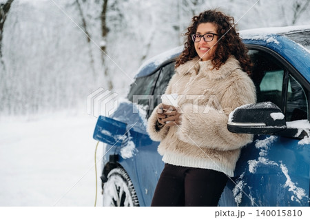 Woman with curly hair in a fur coat stands beside an electric car in a snowy landscape, holding a coffee cup while charging the vehicle during winter Woman with curly hair in a fur coat stands beside an electric car in a snowy landscape, holding a coffee cup while charging the vehicle during winter 140015810