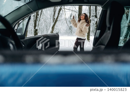 Young girl wearing a fluffy white coat is joyfully playing outside a car in a snowy landscape with trees in the background during winter season Young girl wearing a fluffy white coat is joyfully playing outside a car in a snowy landscape with trees in the background during winter season 140015823
