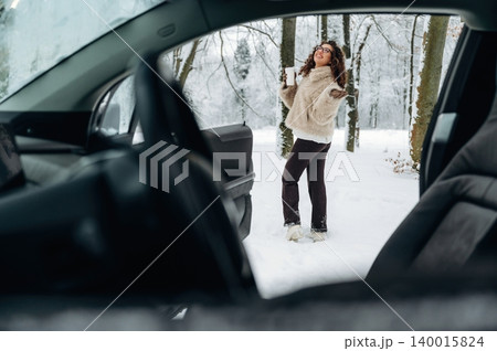 Young woman in a cozy white fur jacket stands outside a car in a snowy forest, surrounded by trees and winter scenery with snow-covered ground and a serene atmosphere 140015824