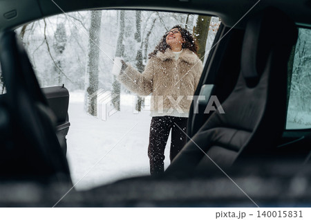 Woman with curly hair wearing a fur coat is enjoying snowfall outside a car in a winter landscape with trees and snow-covered ground visible in the background Woman with curly hair wearing a fur coat is enjoying snowfall outside a car in a winter landscape with trees and snow-covered ground visible in the background 140015831