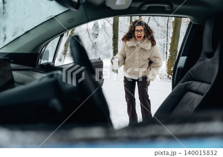 Woman with curly hair wearing a fluffy coat is standing outside a car in a snowy landscape, holding a cup while smiling during a winter day with trees in the background Woman with curly hair wearing a fluffy coat is standing outside a car in a snowy landscape, holding a cup while smiling during a winter day with trees in the background 140015832