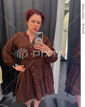 Woman takes a mirror selfie while wearing a brown embroidered dress in a fitting room. Fashion shopping, garment fitting, personal style, retail choice, and everyday wardrobe decision. Woman takes a mirror selfie while wearing a brown embroidered dress in a fitting room. Fashion shopping, garment fitting, personal style, retail choice, and everyday wardrobe decision. 140015888