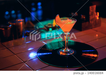 Orange coupe cocktail on vinyl record with dark neon nightclub atmosphere. Orange coupe cocktail on vinyl record with dark neon nightclub atmosphere. 140016084