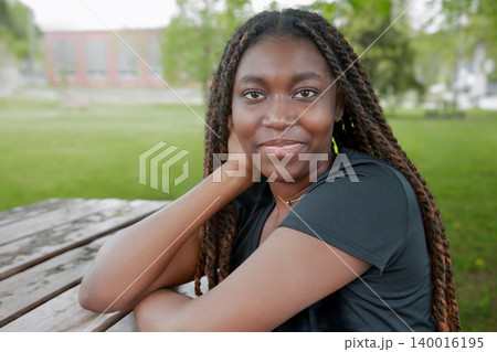 Portrait of a smiling young Black woman with long braids resting at a wooden picnic table in a green park Portrait of a smiling young Black woman with long braids resting at a wooden picnic table in a green park 140016195