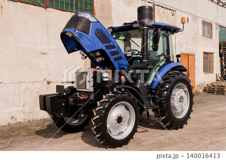 Blue Agricultural Tractor with Open Hood Parked in Front of Old Concrete Farm Building Blue Agricultural Tractor with Open Hood Parked in Front of Old Concrete Farm Building 140016413