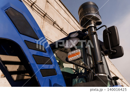 Close up of blue tractor cab with rearview mirrors and headlights against industrial building background 140016425