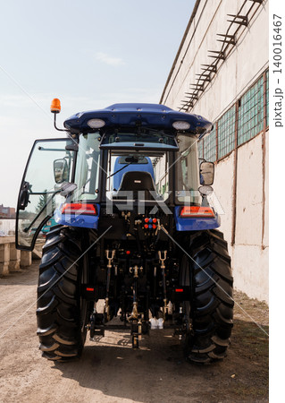 Rear View of Blue Agricultural Tractor Parked Near Industrial Warehouse Building in Daylight Rear View of Blue Agricultural Tractor Parked Near Industrial Warehouse Building in Daylight 140016467
