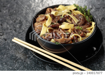 Shimeji and shiitake mushrooms stir-fried with soy sauce, garlic and thyme, served with noodles in a black ceramic bowl. Shimeji and shiitake mushrooms stir-fried with soy sauce, garlic and thyme, served with noodles in a black ceramic bowl. 140017877