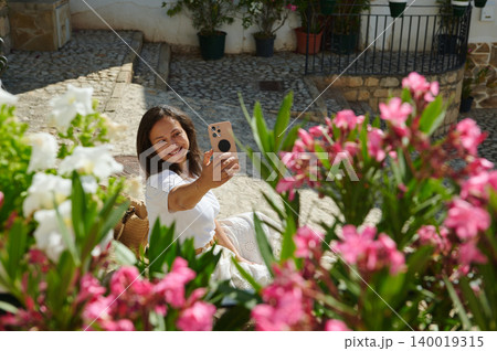 Woman traveling in a small Spanish village taking a selfie among flowers on a cobblestone street Woman traveling in a small Spanish village taking a selfie among flowers on a cobblestone street 140019315