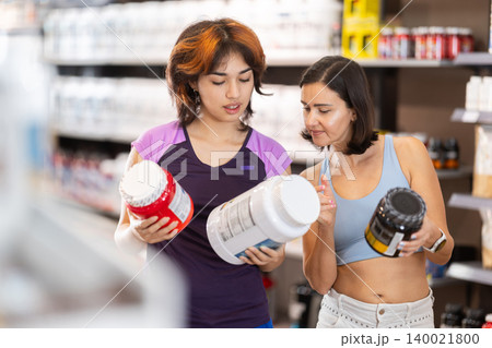 Two woman choosing protein powder jar in sports nutrition store Two woman choosing protein powder jar in sports nutrition store 140021800