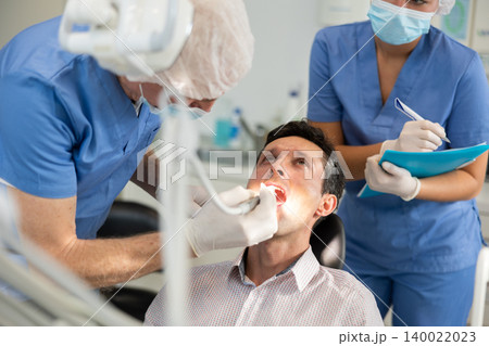 Elderly male dentist cleaning teeth of patient, nurse taking notes Elderly male dentist cleaning teeth of patient, nurse taking notes 140022023