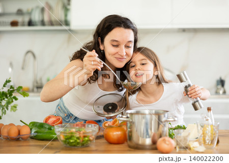 Mom and daughter taste soup in kitchen, take out portion of food from pan. Mom and daughter taste soup in kitchen, take out portion of food from pan. 140022200