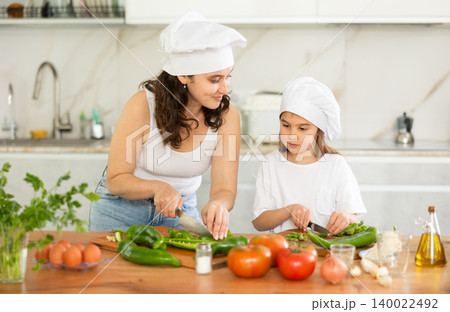 Mom and daughter in hats dice up raw vegetables for salad on cutting boards. Mom and daughter in hats dice up raw vegetables for salad on cutting boards. 140022492