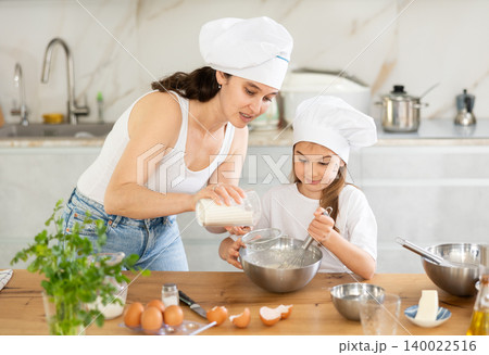 Happy mother and her daughter adding flour in metal bowl and whisking ingredients in the kitchen 140022516